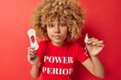 Horizontal shot of curly haired woman holds menstruation sanitary napkin and tampon uses absorbency product dressed in casual t shirt isolated over red background. Periods and menses concept