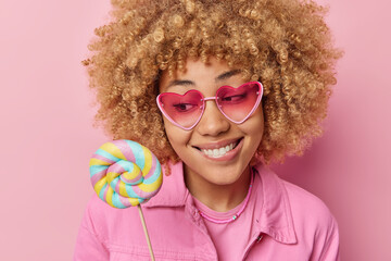 Wall Mural - Pretty woman with curly hair bites lips holds round lollipop has sweet tooth wears trendy sunglasses and jacket isolated over pink background. Positive female model looks at delicious candy.