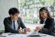 © crizzystudio - Two beautiful Asian businessmen working together in the office sitting at desk with a laptop working on paperwork to discuss the project's financial reports. business organization cooperation concept