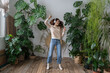 © DimaBerlin - Positive young italian woman in wireless headphones listening to music with pleasure, enjoying the moment, relaxing, dancing on wooden floor in cozy home garden with monstera and tropical plants.