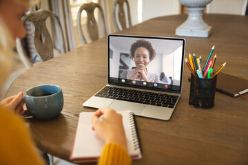 Poster - Caucasian businesswoman writing in notepad while video conferencing with colleague over laptop