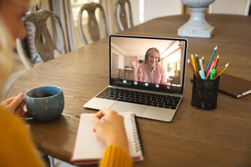 Poster - Caucasian businesswoman writing in notepad while video conferencing with teammate over laptop