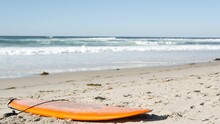 Surfboard For Surfing Lying On Beach Sand, California Coast, USA. Ocean Waves And Orange Surf Board Or Paddleboard. Longboard Or Sup For Watersport By Sea Water. Summer Vacations, Sport On Shore Vibes