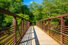 A Long Metal Rust Colored Bridge Over The Silky Brown Waters Of Little River Surrounded By Lush Lush Green Trees, Grass And Plants With Blue Sky And Clouds At Olde Rope Mill Park In Woodstock Georgia 