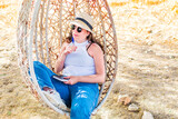 Fototapeta Natura - Woman sitting in hanging rattan egg chair, relaxing summer.
