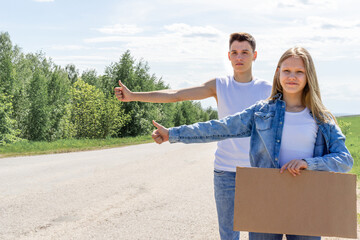 A couple of teenagers are trying to stop a car on the road with an empty cardboard sign, a mock-up. Car travel. Young people are hitchhiking. Two young people tourists hitchhiking on the road. High