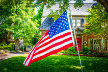 Waving American Flag Large  In Front Of Blurred Upscale Two Story Landscaped Home With Car Behind Security Gate Surrounded By Shady Leafy Trees