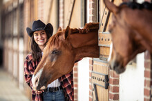 Two Horses And A Young Woman In Cowboy Hat Next To Each Other In The Stables