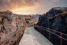 Tourists Walking On Metallic Pedestrian Bridge. Built Structure Amidst Rocky Cliffs Leading Mountain Against Cloudy Sky. People Enjoying At Thingvellir National Park During Sunset.