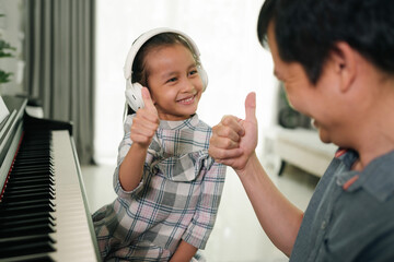 Wall Mural - Asian daughter and father showing thumb and smiling together while playing piano at home, concept of love, bonding, relation, education, music, skill, mental health, parent and child in family life.