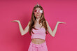 Surprised young woman in funky crown keeping arms outstretched while standing against pink background