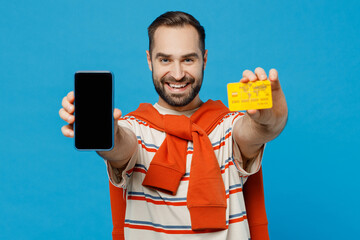 Wall Mural - Young man in orange striped t-shirt using mobile cell phone with blank screen workspace area hold credit bank card doing online shopping order delivery booking tour isolated on plain blue background