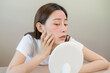 © KMPZZZ - Dermatology, puberty asian young woman, girl looking into mirror, allergy presenting an allergic reaction from cosmetic, red spot or  rash on face. Beauty care from skin problem by medical treatment.