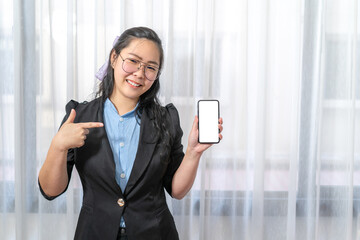 Wall Mural - Happy Asian woman fingering up and pointing with mobile phone white screen with her hand at copy space while standing in front of the window.