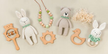 Infant Toys On A Pastel Beige Background. Wooden And Crocketed Baby Handmade Toys For A Banner In Eco Style. Baby Toys Concept. Wooden Rattles, Knitted Teddy Bear And Teething Beads Top View.