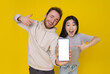 © Svyatoslav Lypynskyy - Caucasian guy and asian girl show thumbs up excited holding smartphone with blank white screen, mobile app advertisement isolated on yellow background. Product placement. Great offer