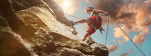 Muscular Climber Man In Protective Helmet Abseiling From Cliff Rock Wall Using Rope Belay Device And Climbing Harness On Evening Sunset Sky Background. Active Extreme Sports Time Spending Concept.