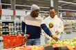 © pressmaster - Young man with shopping cart pointing at fresh apples while helping his girlfriend choose fruit in grocery department of supermarket