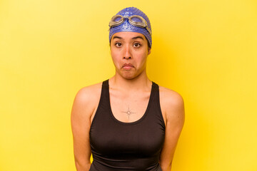 Wall Mural - Young swimmer hispanic woman isolated on yellow background shrugs shoulders and open eyes confused.