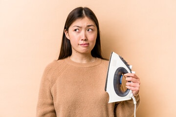 Wall Mural - Young asian woman holding iron isolated on beige background confused, feels doubtful and unsure.