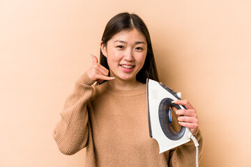 Wall Mural - Young asian woman holding iron isolated on beige background showing a mobile phone call gesture with fingers.