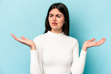 Wall Mural - Young caucasian woman isolated on blue background confused and doubtful shrugging shoulders to hold a copy space.