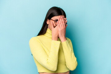 Wall Mural - Young caucasian woman isolated on blue background blink at the camera through fingers, embarrassed covering face.