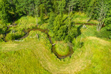 Fototapeta  - Mała wąska rzeka płynąca zakolami przez las. Brzegi porasta zielona trawa, w głębi znajduje się mieszany las. Jest słoneczny dzień. Zdjęcie zrobione z wysokości przy użyciu drona.