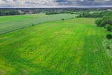 Fototapeta Las - Rozległy, płaski teren pokryty zielonymi polami uprawnymi i łąkami. Widać polną drogę, kępy drzew, w oddali zabudowania pobliskiej miejscowości. Widok z wysokości, zdjęcie zrobione z użyciem drona.