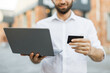 © sofiko14 - Focus on laptop and bank card. Business trip concept. Handsome young man, using notebook and holding credit card for shopping online. Male freelancer receive payment online.
