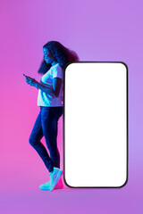 Poster - Full length of young black lady using mobile device, standing near huge cellphone with mockup for design in neon light