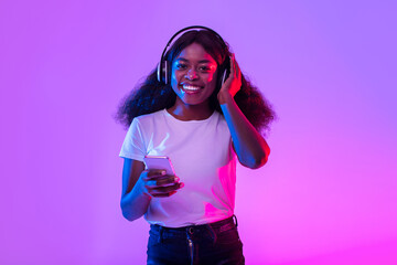 Poster - Happy millennial black female in wireless headphones using smartphone in neon light