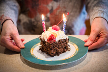 Close Up Of Woman Holding A Plate With A Slice Of Birthday Cake With Lit Candles. Piece Of Black Forest Cake And Small Serving Size. Birthday Party Food And Desserts Background 