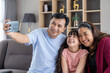 © amorn - Happy Asian family with teen child daughter having fun taking selfie on mobile smartphone at home. Smiling parents and teenage kid girl embrancing and posing for take family photo on sofa.
