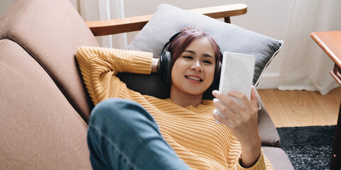 Close up smiling attractive woman in headphones using smartphone, lying on couch, positive young female looking at phone screen, watching video or making call, chatting online, listening music