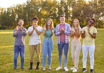 Cheerful young female and male friends applaud together during summer walk in park. Smiling multiracial friends in casual clothes stand in row and clap their hands. Concept of friendship and unity.