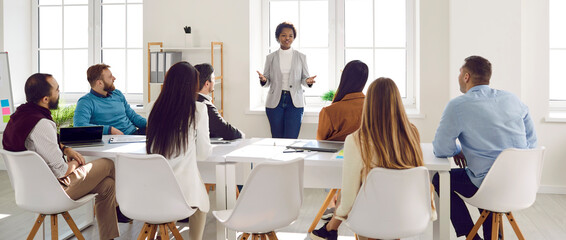 Young business people sitting around big table and listening to team leader. Black woman standing in modern white office boardroom during meeting and speaking to group of multiracial colleagues