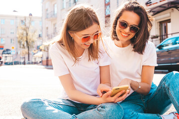 Wall Mural - Two young beautiful smiling hipster female in trendy summer white t-shirt clothes and jeans.Sexy carefree women posing on the street background.Positive models using smartphone apps, looking at screen