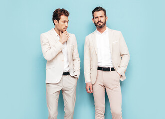 Portrait of two handsome confident stylish hipster lambersexual models. Sexy modern men dressed in white elegant suit. Fashion male posing in studio near blue wall