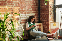 Young Woman With Dark Long Wavy Hair Holding Retro Photocamera In Hands While Sitting Against Brick Wall In Spacious Loft Apartment