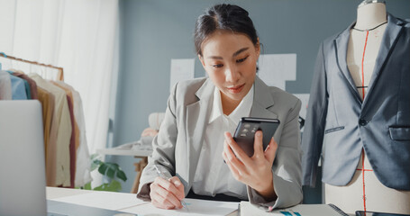 Wall Mural - Professional cheerful Asia businesswoman designer sitting on table  look at smartphone check purchase order online marketplace with stock inventory on laptop computer at studio workplace.