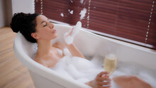 Young Playful Woman Relaxing In Hot Bath With Bubbles Holding Glass Champagne In Hand Enjoying Luxury Bathroom Romantic Happy Girl Taking Bathtub Plays Blowing On Foam Drinking Alcoholic Drink Resting