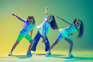 Group of children, little girls in sportive casual style clothes dancing in choreography class isolated on green background in yellow neon light. Concept of music, fashion, art