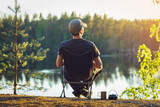 Fototapeta  - A man is sitting in a camping chair on a summer evening on the background of a forest lake.