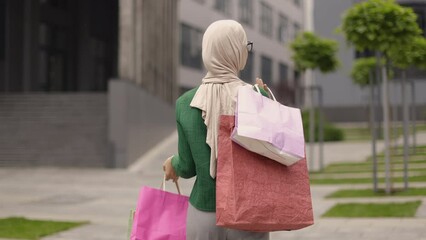 Wall Mural - Close up portrait of beautiful young muslim woman, wearing casual clothes, going for shopping in background of modern city buildings, posing to camera with shopping bags. Copy space.