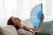 Relaxed tired young european woman with closed eyes suffering from heat on sofa and waving fan at herself