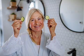 Poster - Beautiful senior woman in bathrobe applying cucumber face mask in bathroom, skin care concept.