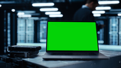 Poster - Laptop Computer with Green Screen Chroma Key Mock Up Display Stands on a Table. In the Background Data Center With Rows of Server Racks and IT Engineer Working. Advanced Equipment.