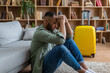 © Prostock-studio - Vacation cancellation concept. Upset black man sitting near suitcase at home and touching forehead, waiting for flight