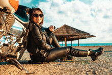 Independent Happy Woman In A Leather Motorcycle Outfit Poses Sitting On The Ground Near A Motorcycle. Ocean And Beach Umbrellas On The Background. The Concept Of The Motorcycle Travel And Feminism